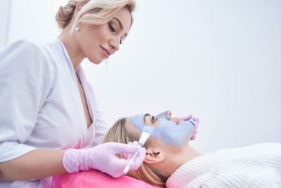 Cosmetician spreading blue clay paste over facial skin of client using two brushes